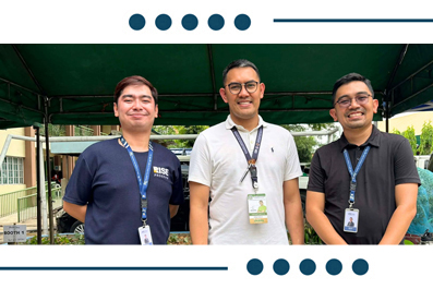 Three men with lanyards stand together at an outdoor event under a green tent, posing for a photo.