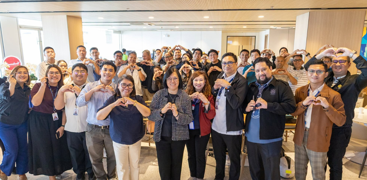 Large group of colleagues indoors, many forming heart shapes with their hands for a team event or celebration.