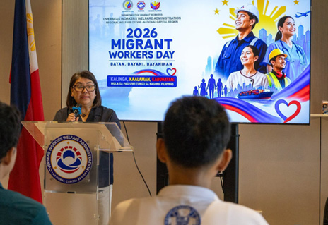 Woman speaks at a lectern during the 2026 Migrant Workers Day event, with a banner showing migrant workers and a heart logo in the background.