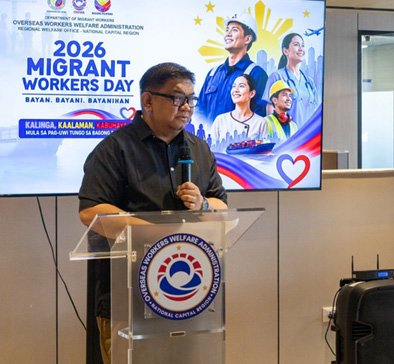 Man in a dark shirt speaks at a transparent podium during the 2026 Migrant Workers Day event, with a banner and workers' illustration on a screen behind him.