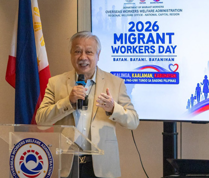 Senior man in a beige blazer speaks into a microphone at a clear lectern during 2026 Migrant Workers Day, with the Philippine flag nearby and a conference banner behind him.
