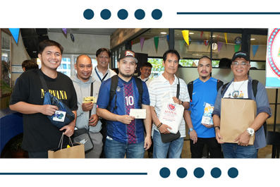 Eight men stand together at a festive event, posing with certificates, tickets and gift bags under colorful banners.