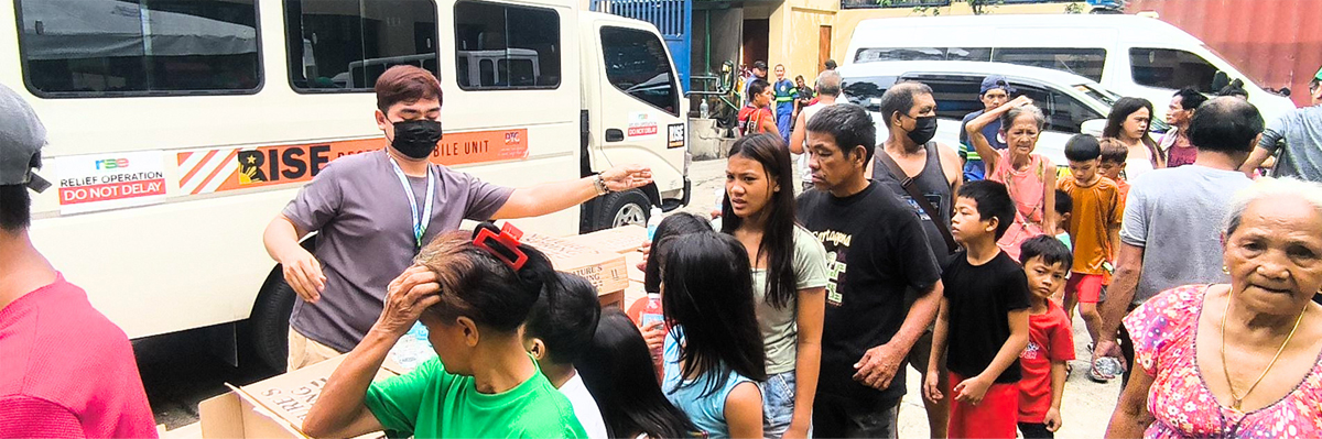People line up for aid beside a relief truck labeled 'Relief Operation' with a masked man directing the queue and boxes in view.