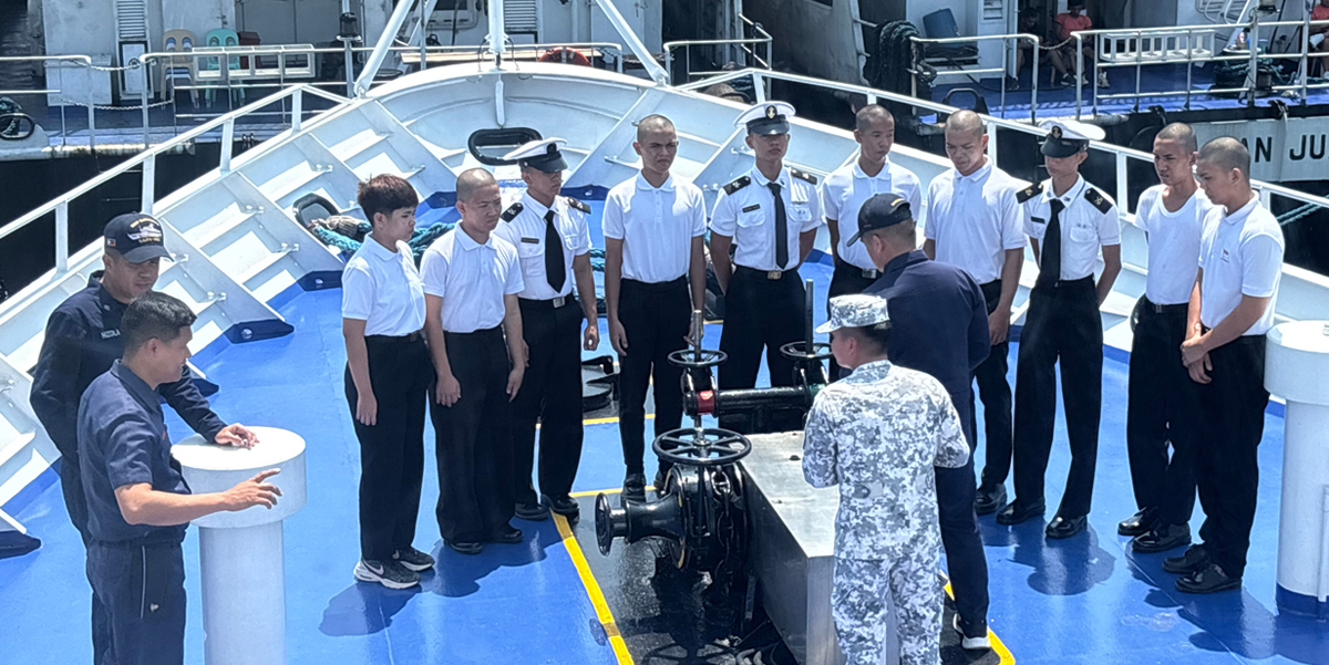 Navy trainees in white shirts form a semicircle around a large valve on a blue ship deck, listening to an instructor in camouflage.