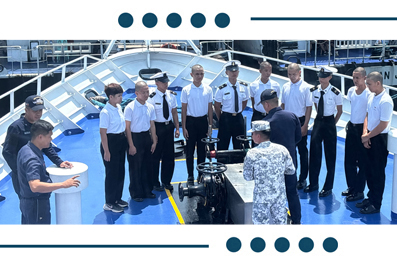 Group of naval officers and sailors on a ship deck listening to a presenter beside a large black marine equipment on the blue deck.