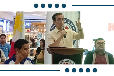 Man at a podium giving a speech with a microphone, Philippine flag visible on the left and attendees nearby as part of a public event.