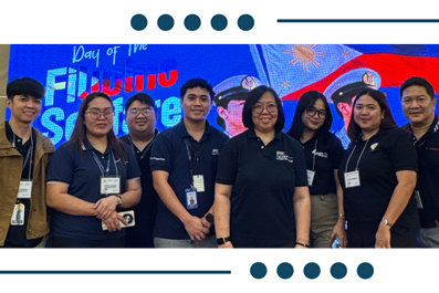 Group of eight staff members in navy polo shirts with lanyards posing for a photo at a booth, blue banner backdrop visible behind them.