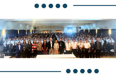 Large group of people posing for a group photo in a conference hall, with rows of attendees standing behind a table on stage.