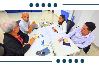 Four professionals in a meeting around a table, reviewing a large document; coffee cups, notebooks and a smartphone on the table.