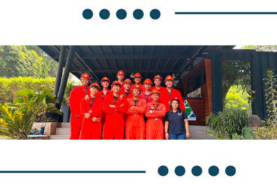 Group of workers in red uniforms and hard hats posing for a team photo in front of a wooden building, with one person in a blue shirt on the right.