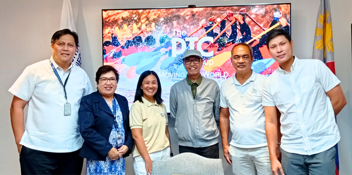 Six people pose for a group photo indoors, in front of a colorful screen that reads 'The PTC Up Moving World', with flags on both sides.