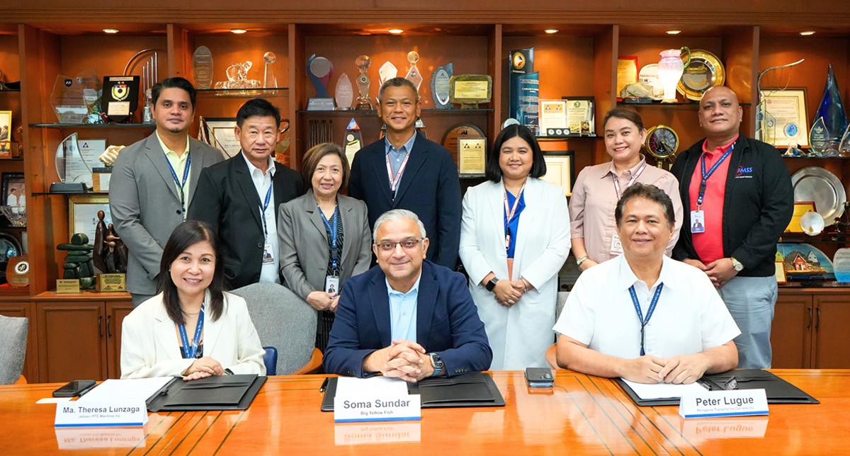 PTC JPMI and Big Yellow Fish 02 | PTC Group of professionals posing for a formal photo in a boardroom; three people sit at a conference table with name plates—Ma. Theresa Lunzaga, Soma Sundar, and Peter Lugue—while colleagues stand behind them amid awards on shelves.