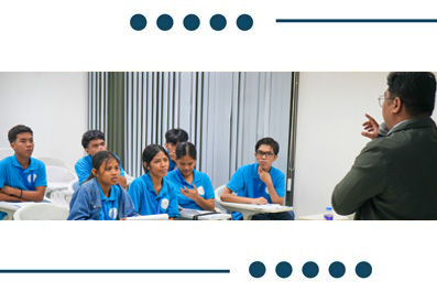 Teacher leads a discussion at the front of a classroom with students in blue uniforms seated at desks.