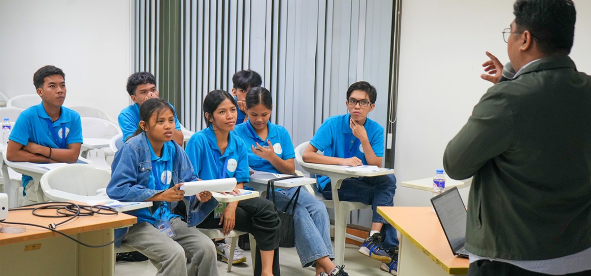 PTC welcomes PIC participants 02 | PTC Group of students in blue shirts seated in a classroom, listening to a presenter at the front with a laptop and papers on a desk.