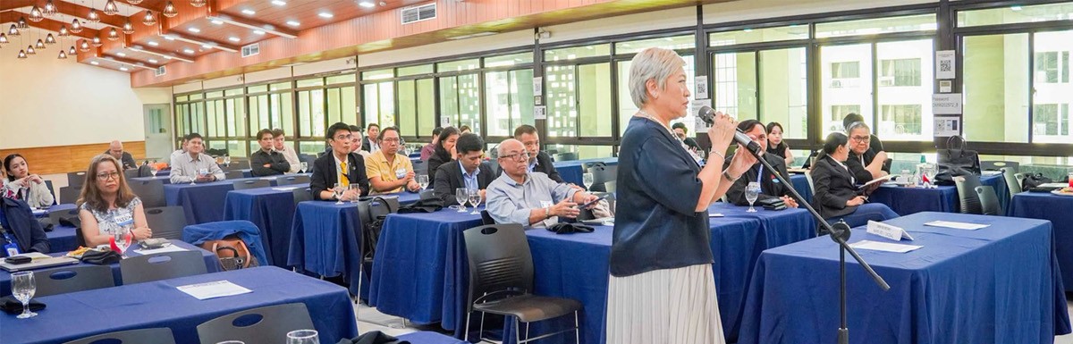 PTCs Karen Avelino champions 03 | PTC An older woman speaks into a microphone at a podium during a conference, with attendees seated at blue-draped tables.