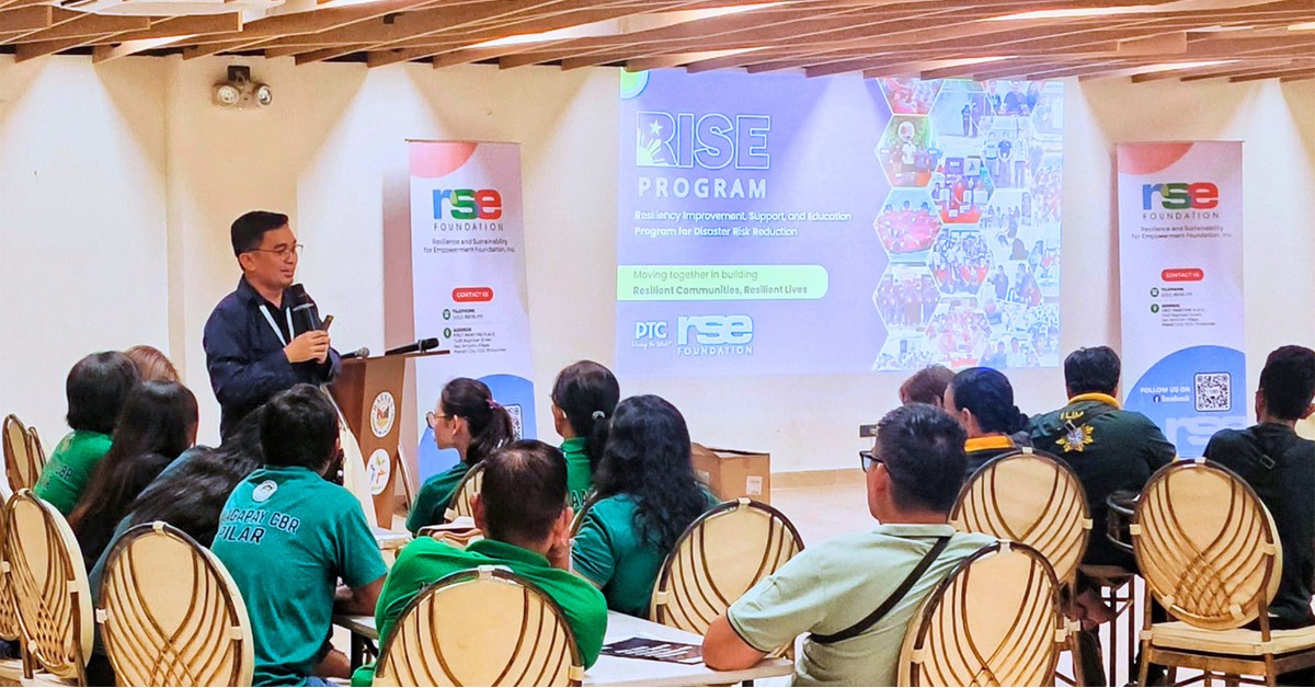 RSE Foundation facilitates RISE Citizen 02 | PTC Male presenter at a podium speaks to an attentive audience in a conference room, with a large RISE Program projection and banners nearby.