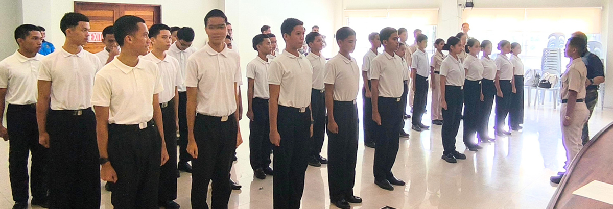 Group of young cadets in white shirts and dark pants standing in formation as an instructor in a tan uniform speaks to them in a bright room