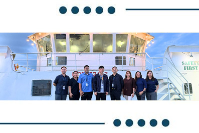 Eight people pose for a team photo on a ship's deck with a cockpit window behind them and a 'Safety First' sign visible.